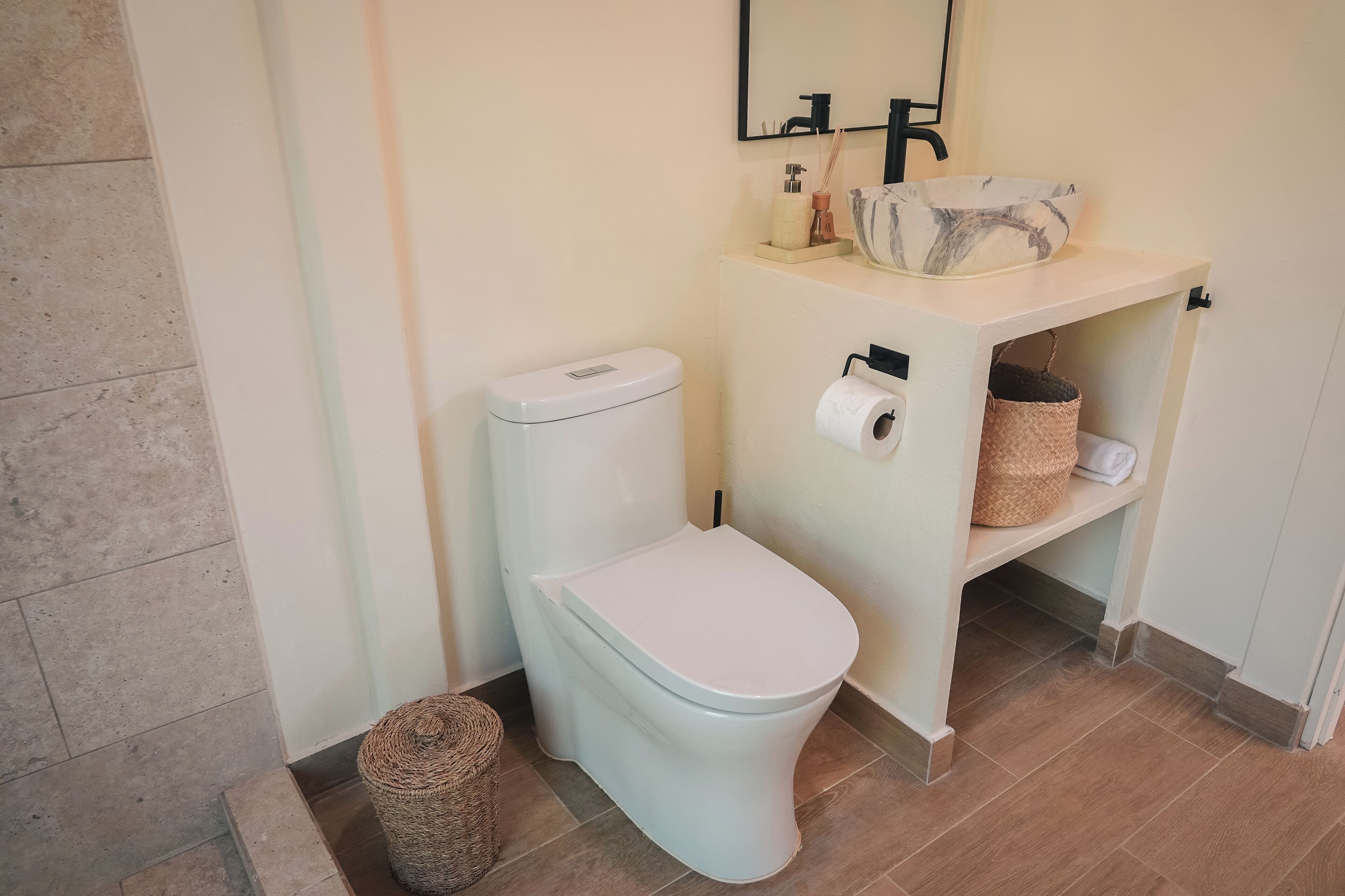 Un baño moderno con inodoro blanco, lavabo de mármol y estante con artículos de aseo.
