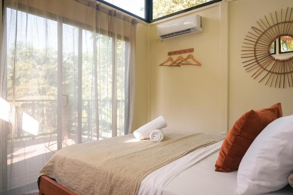A bedroom with a bed, a window with sheer curtains, a wall-mounted air conditioner, and a decorative mirror.