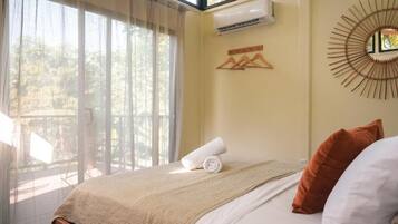 A bedroom with a bed, a window with sheer curtains, a wall-mounted air conditioner, and a decorative mirror.