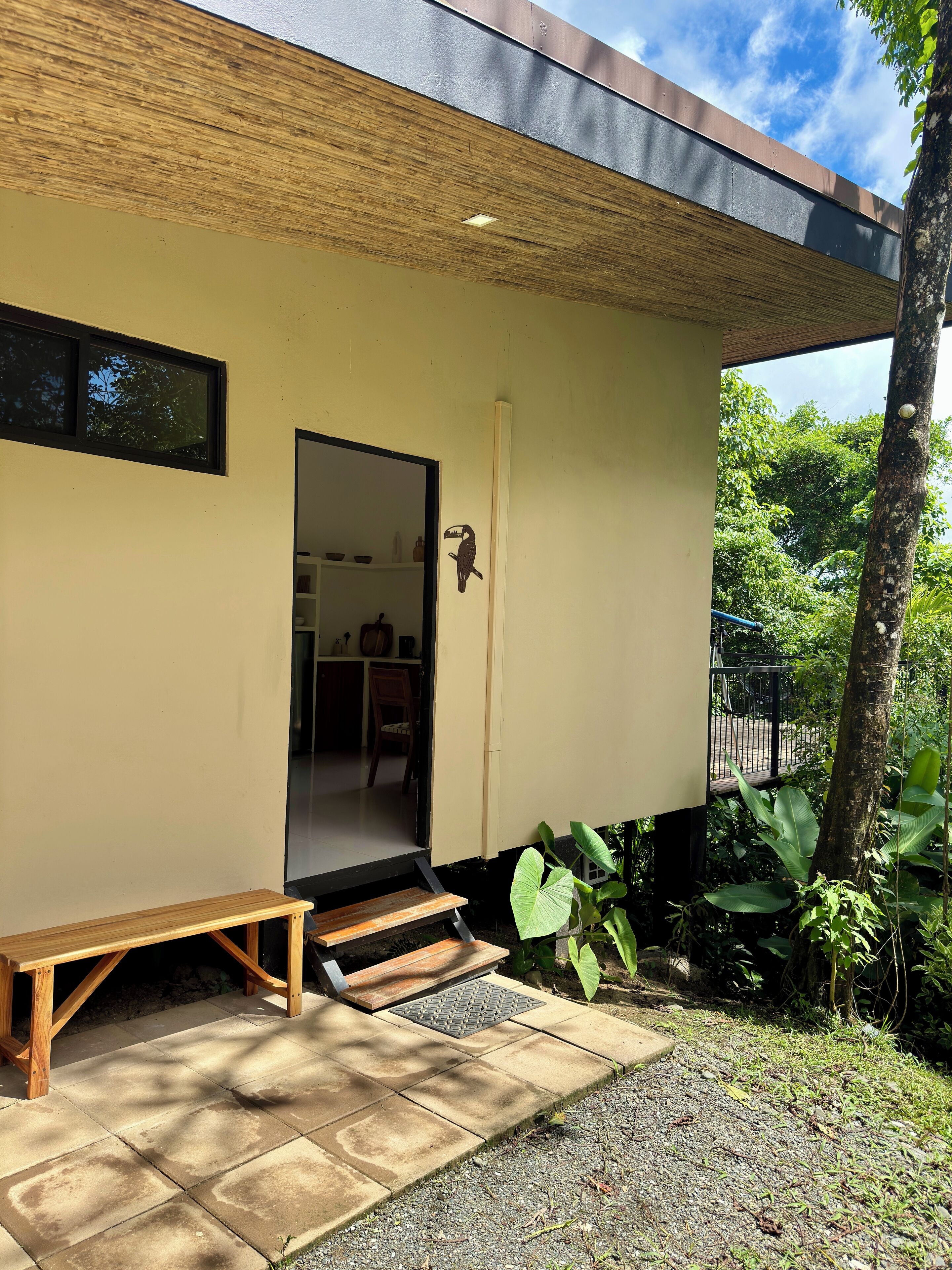 Une maison moderne avec un banc en bois à l’extérieur, une porte donnant sur l’intérieur et un jardin agrémenté d’un arbre et de plantes.