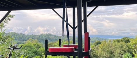 An outdoor gym with exercise equipment, including a bench press and a pull-up bar, set against a backdrop of lush greenery and a mountainous landscape.
