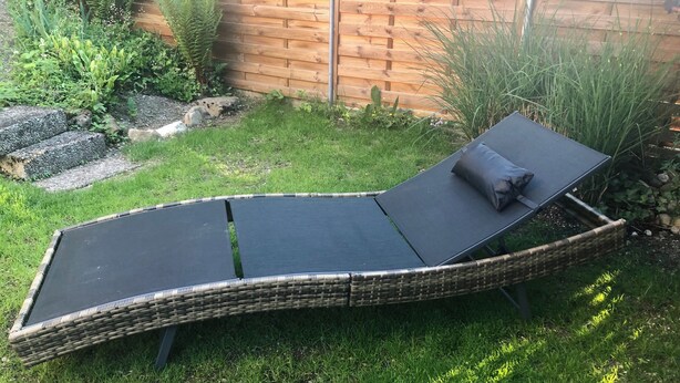 A wicker lounge chair with a black cushion and a matching pillow, positioned on a grassy lawn with a wooden fence and plants in the background. - Apartment Deluxe (Rapperswil-Jona)