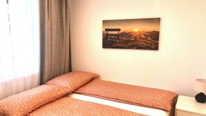A room with two beds, a window with curtains, a wall-mounted picture of a sunset, and a bedside table with a lamp. - Apartment Deluxe (Rapperswil-Jona)