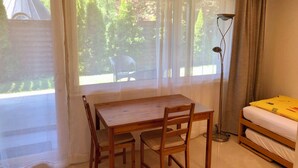 A room with a wooden table and chairs, a bed with a yellow cover, a floor lamp, and a view of greenery outside. - Apartment Deluxe (Rapperswil-Jona)