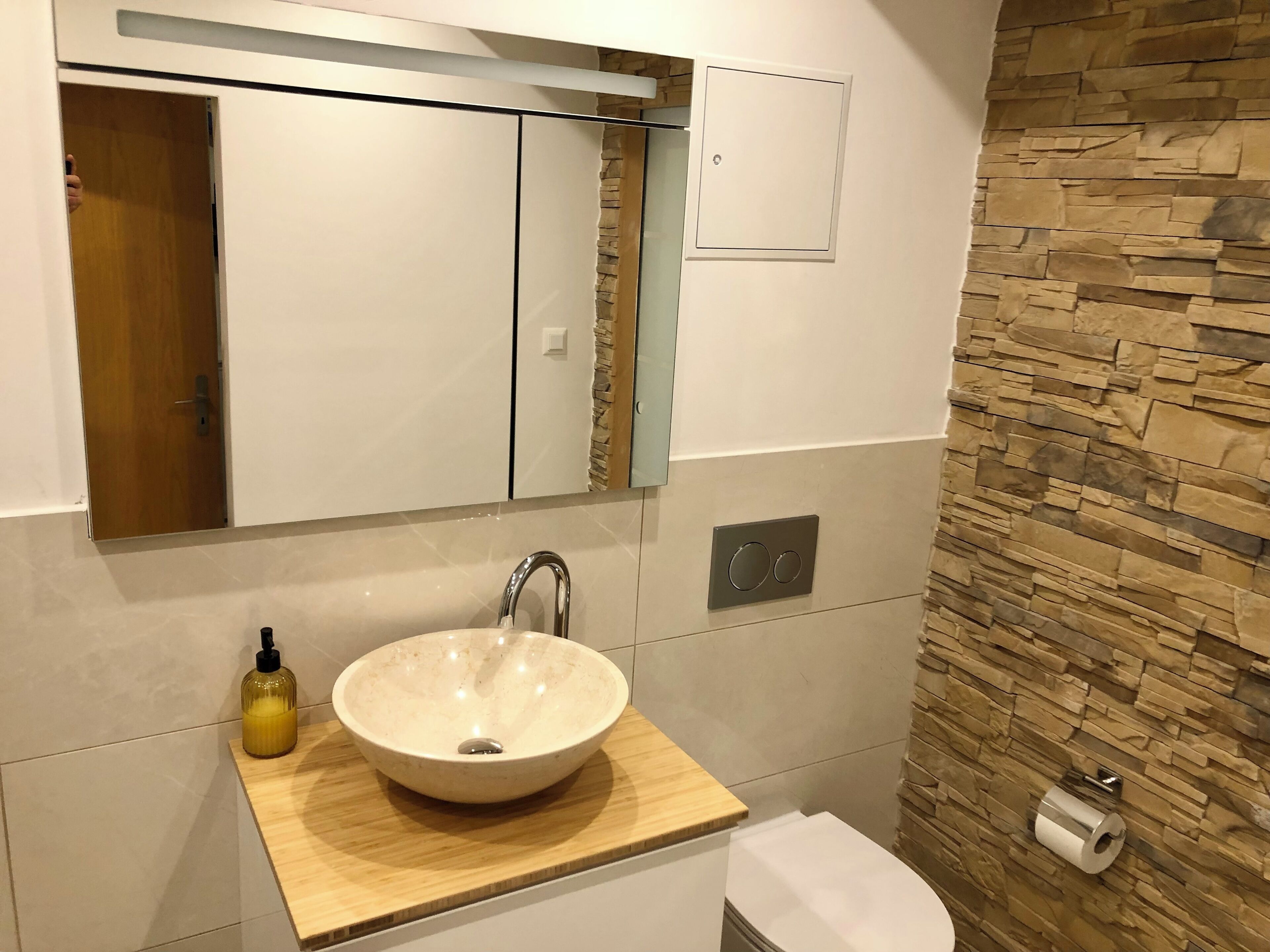 A modern bathroom with a wooden vanity, a large mirror, a white sink, and a stone wall.