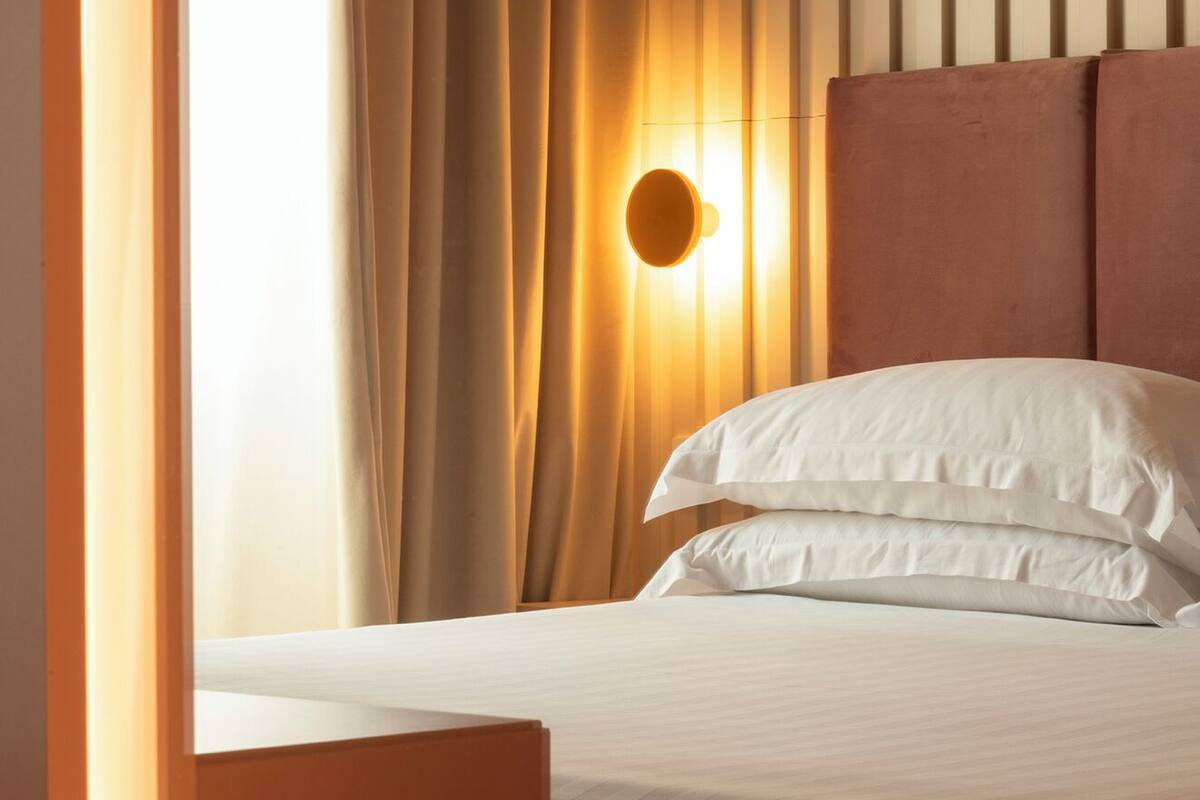 Ein ordentlich bezogenes Bett mit weißen Leinentüchern, ein Nachttisch aus Holz und eine Wandlampe.