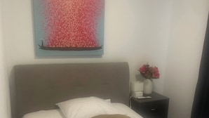 A neatly made bed with white linens and a brown pillow, a small bedside table with a vase of flowers, and a painting of a boat on a body of water on the wall. - Anita Hostel (Zürich)