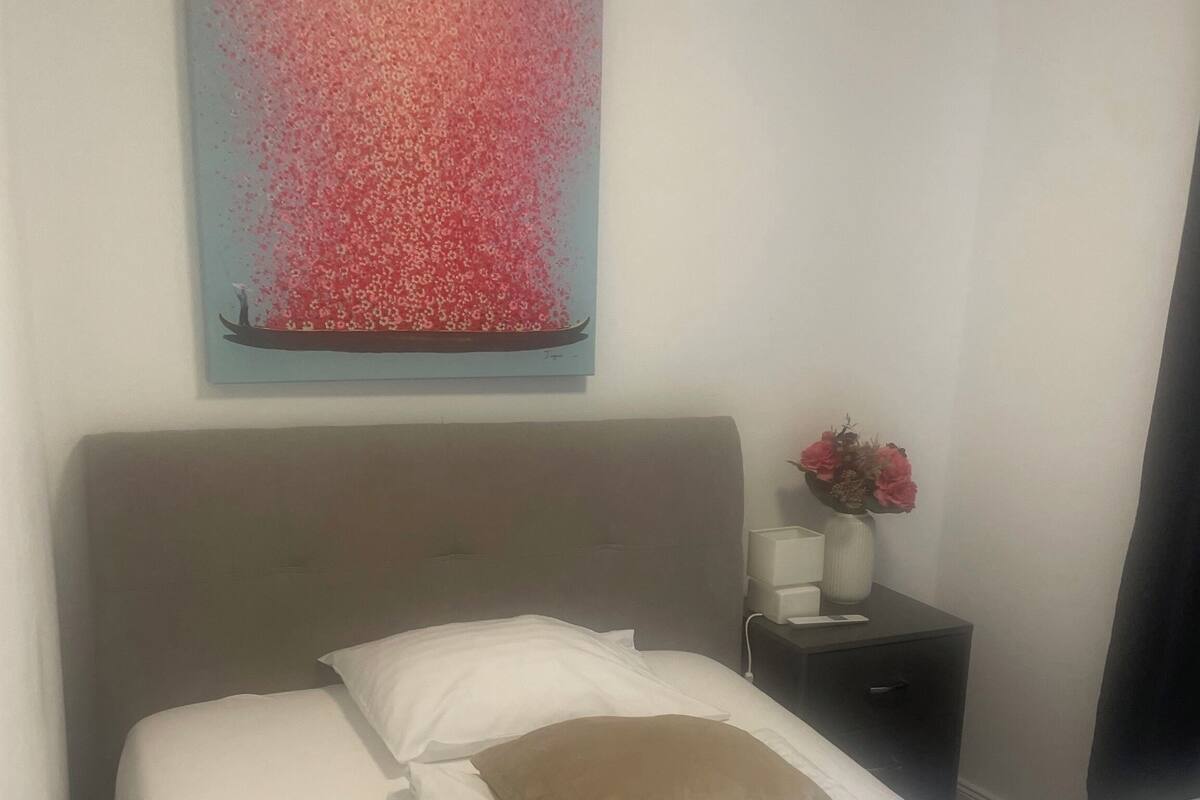 A neatly made bed with white linens and a brown pillow, a small bedside table with a vase of flowers, and a painting of a boat on a body of water on the wall.