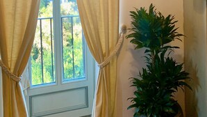Et værelse med et stort vindue, gardiner, en potteplante, et indrammet billede og en reol med en vase.