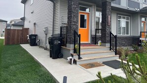 A two-story house with a grey exterior, a stone accent wall, and an orange front door. There are two black trash bins and a solar panel on the front porch. - Stylish New Home, Near Shops, WiFi, 2V Garage, Smart TV, Private Yard, Near ✈️. (Edmonton)