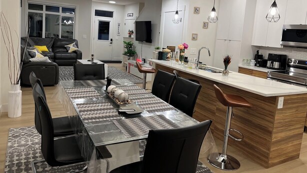 A modern kitchen with a central island, bar stools, and a dining area with a glass table and chairs. - Stylish New Home, Near Shops, WiFi, 2V Garage, Smart TV, Private Yard, Near ✈️. (Edmonton)