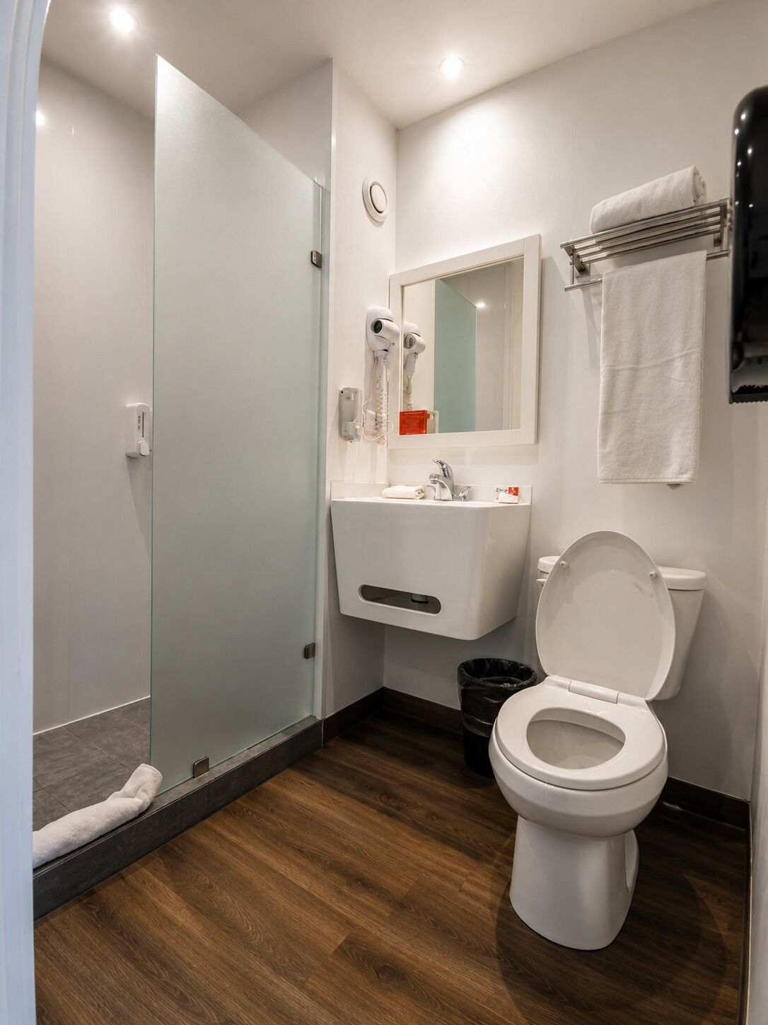 Un baño moderno con puerta de ducha de vidrio, inodoro blanco, lavamanos con espejo y toallero.