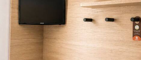 A small room with a mounted television, a wooden shelf, a stool, and a trash bin.
