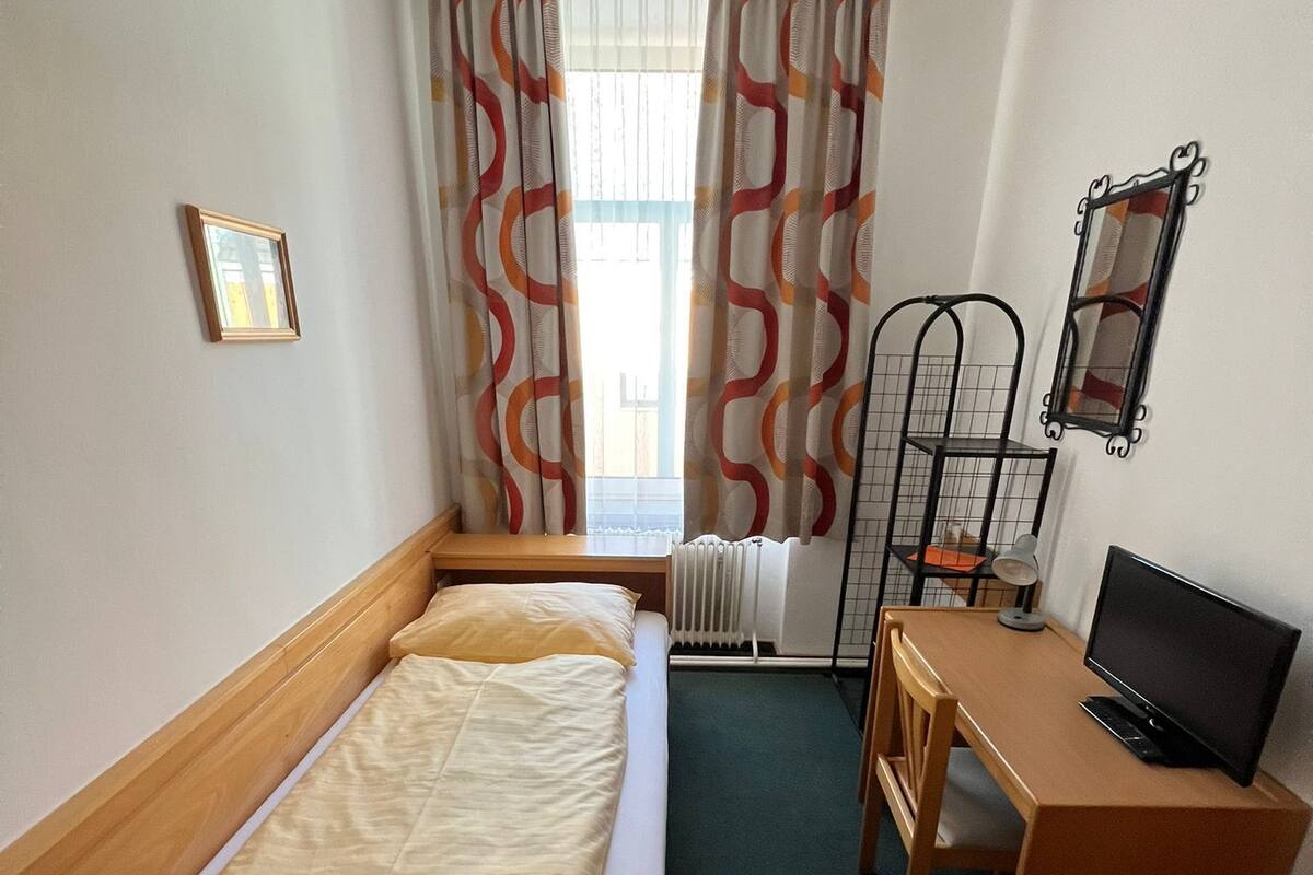 Une chambre d’hôtel de taille réduite, comprenant un lit simple, un bureau avec un ordinateur et un radiateur.