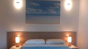 Un dormitorio con cama, dos lámparas de noche, una mesita de noche y un cuadro en la pared con una escena de playa.