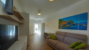 Una sala de estar moderna con un televisor de pantalla plana, un sofá color beige con cojines verdes, piso de madera y un cuadro de una escena costera en la pared.