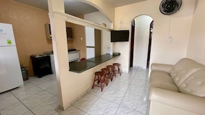 A compact kitchen with a refrigerator, stove, and microwave. There is a bar area with stools and a TV mounted above it. A beige sofa is in the living area.