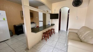 Cocina compacta con refrigerador, estufa y microondas. Hay una barra con taburetes y un televisor montado encima. Un sofá color beige se encuentra en la sala.