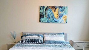 A bedroom with a bed, a nightstand, a drawer unit, and a wall-mounted abstract painting.