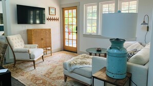 Ein Wohnzimmer mit einem weißen Sofa, einem weißen Sessel, einer blauen Vase, einem Fernseher und einer zur Außenseite führenden Tür.