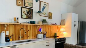 Eine kleine Küche mit Spüle, Herd und weißen Schränken. Ein Korbstuhl und ein Teppich liegen auf dem Boden.