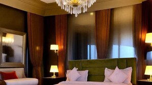 A large bed with a green headboard, white bedding, and multiple pillows. There are two bedside tables with lamps, a large mirror, and dark curtains. - The Rules Club (Mat)