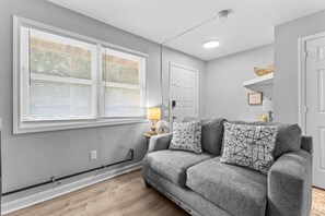 A room with a grey couch, two patterned pillows, a white door, and a window with blinds.