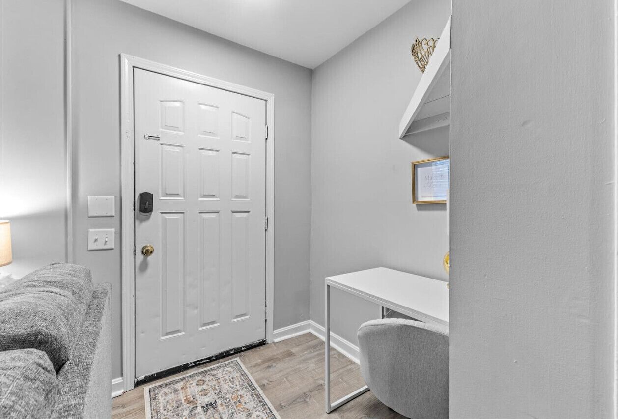 A room with a white door, a gray sofa, a small white desk, and a patterned rug.