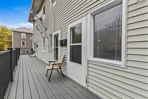 A wooden deck with a chair and a table, a white door with a window, and a mailbox on the side of a house. - Comfortable 2BR in Winooski! (Winooski)
