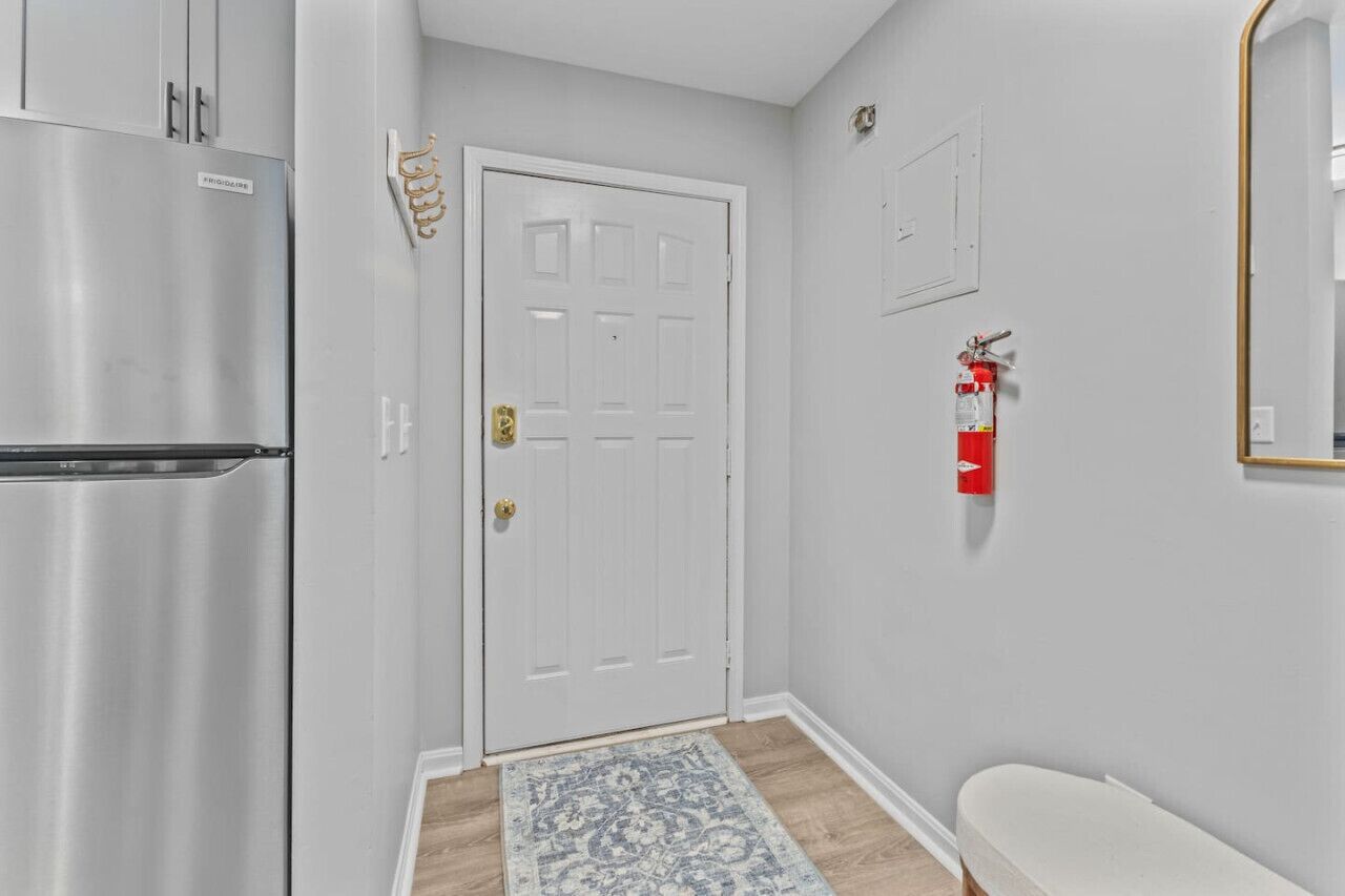 A hallway with a white door, a stainless steel refrigerator, a red fire extinguisher, and a patterned rug.