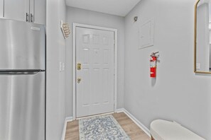 Un pasillo con una puerta blanca, un refrigerador de acero inoxidable, un extintor rojo y una alfombra estampada.