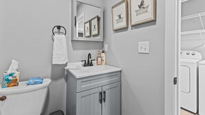Een badkamer met toilet, wastafel en spiegel. Er hangen foto's in lijsten aan de muur, een handdoek hangt aan de muur, en er staat een handzeepfles op de wastafel.