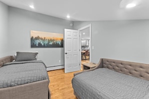 A bedroom with two beds, a painting of a forest, a white door, and a view into a kitchen area. - Unique Top-Floor Apt in BTV (Burlington)