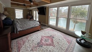 A bedroom with a large bed, a wooden ceiling, a TV, and a view of a lake through large windows. - Family-Friendly Lakefront Home with Hot Tub, Firepit, Dock & Breathtaking Views! (Charlotte)