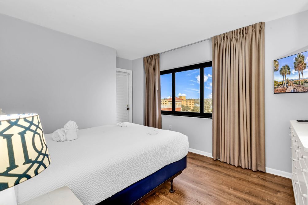 Une chambre d’hôtel avec un grand lit, une fenêtre avec des rideaux, un tableau représentant des palmiers et une commode blanche.