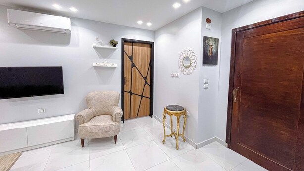 A modern living room with a flat-screen TV, a beige armchair, a wooden door, and a small round table.