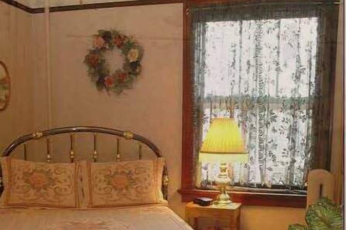 A bedroom with a bed, a nightstand with a lamp, a chair, a window with curtains, and a wall decoration.