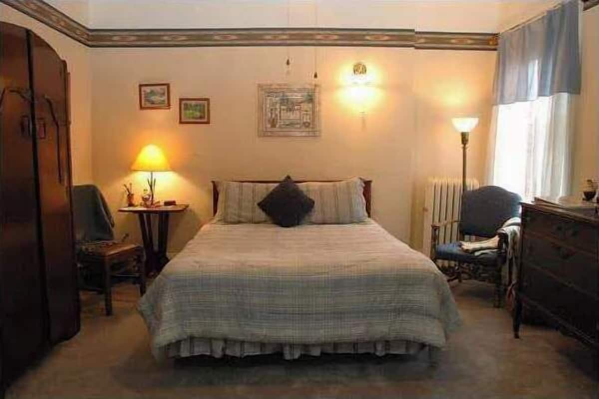A bedroom with a bed, a nightstand, a lamp, a chair, a dresser, and a window with curtains.