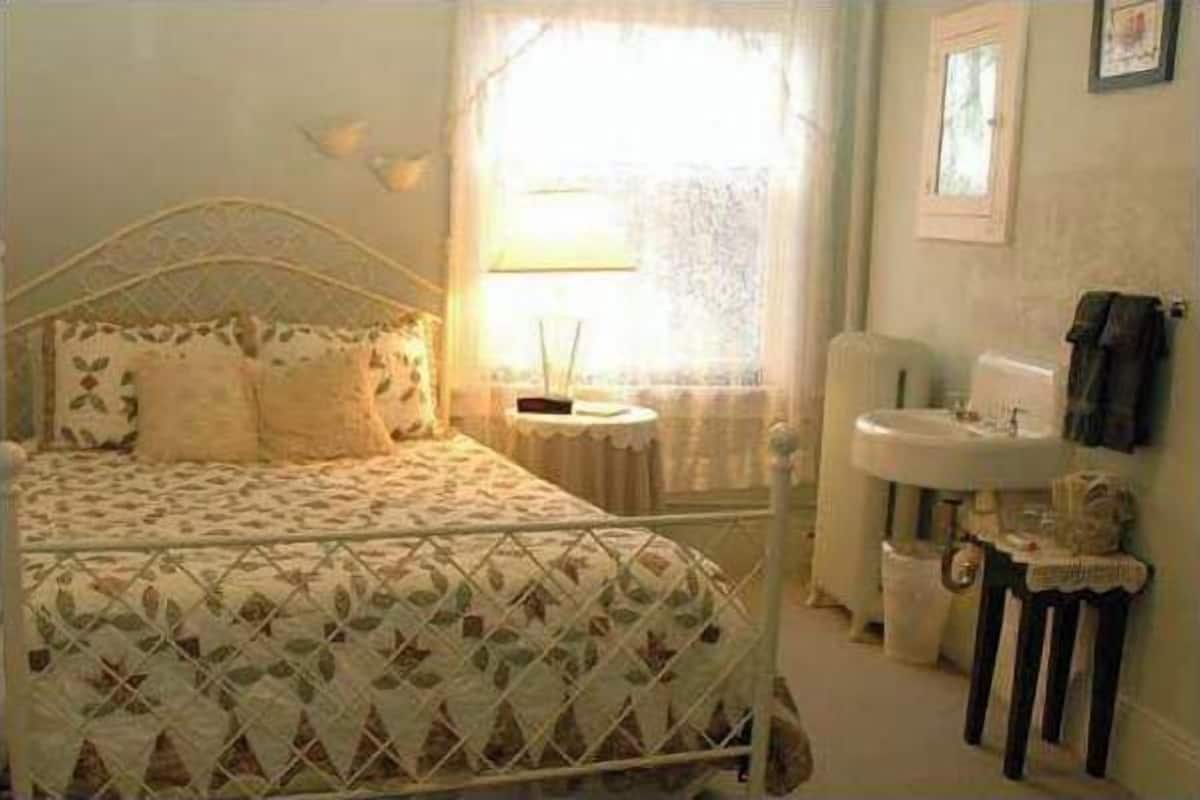 A bedroom with a bed, a window with a lace curtain, a bedside table, and a small white sink.