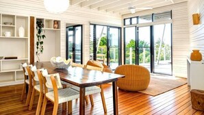 Un espace repas comprenant une table et des chaises en bois, un pouf en fibres tressées et de grandes fenêtres donnant sur un paysage verdoyant.