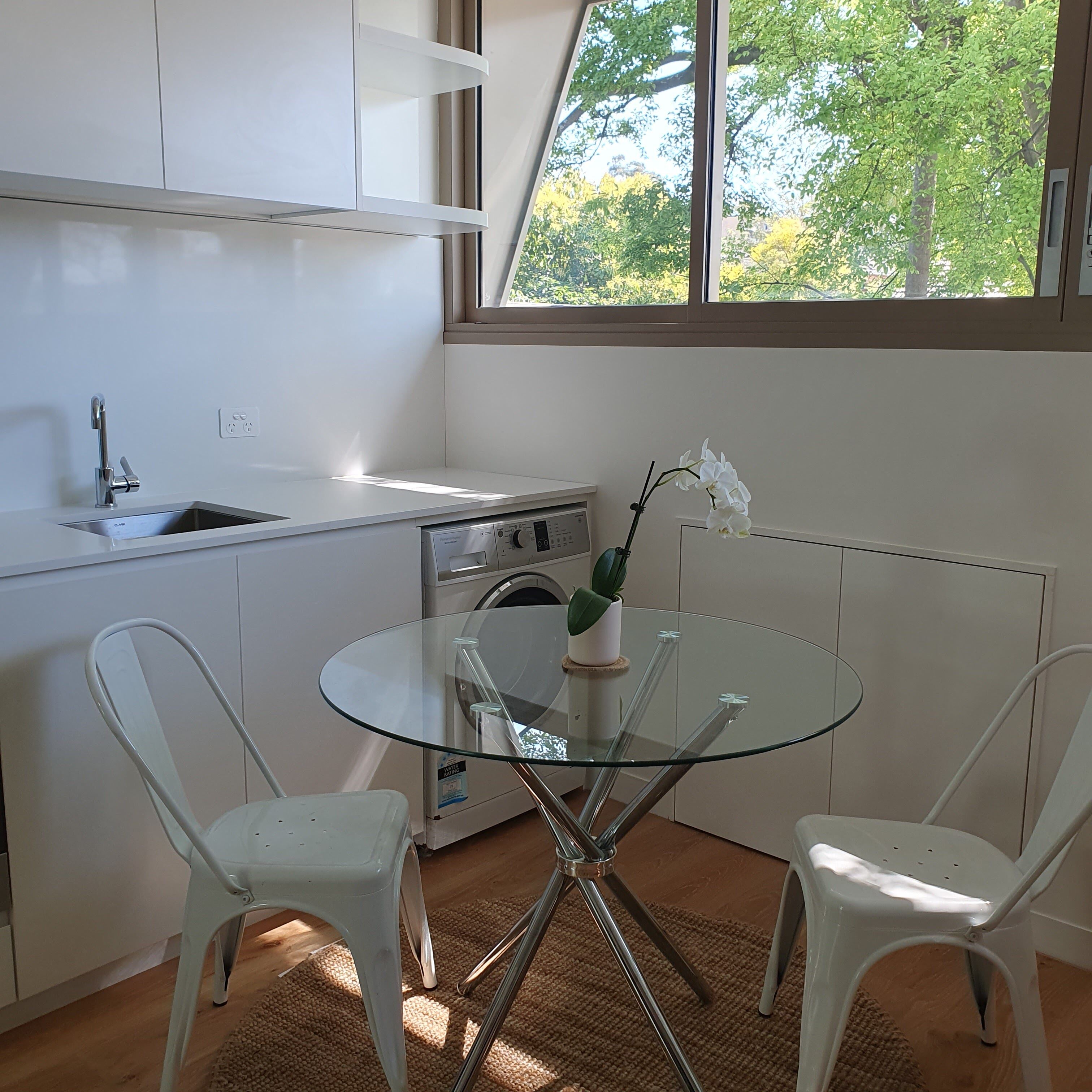 A compact kitchen with a sink, a washing machine, and a glass table with chairs.