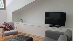 A modern living room with a grey sofa, a black leather ottoman, and a wooden chair with a colorful cushion. A flat-screen TV is mounted on a white wall above a white cabinet. - 1 BR unit with leafy outlook & dedicated workspace (Glebe)