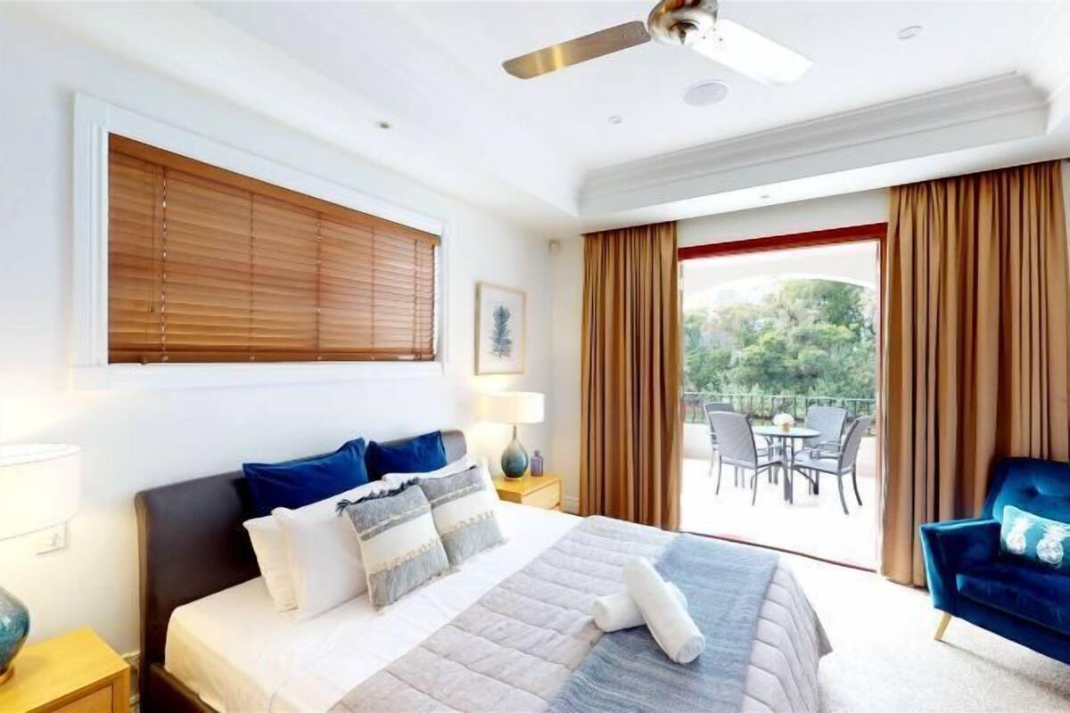 A bedroom with a bed, a ceiling fan, a window with blinds, a balcony with a table and chairs, and a blue armchair.