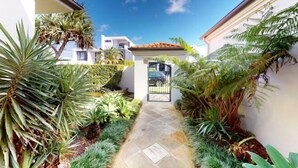 A narrow pathway leading to a gated entrance, surrounded by lush tropical plants and a clear blue sky. - Luxico Lakeland Keys (Heated Pool) (Broadbeach Waters)