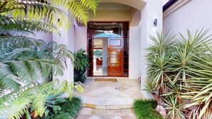 A modern building entrance with a stone pathway, a wooden door, and lush greenery including ferns and palm-like plants. - Luxico Lakeland Keys (Heated Pool) (Broadbeach Waters)