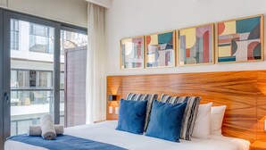 Una habitación de hotel moderna con una cama grande, cabecera de madera y vistas de la ciudad a través de ventanales de piso a techo.