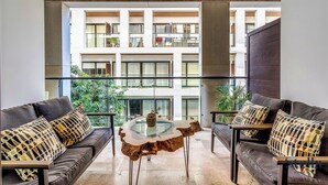 Una sala de estar moderna con una mesa de centro de vidrio, dos sofás con cojines estampados y vistas de un edificio de varios pisos con balcones.