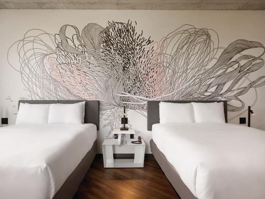 A hotel room with two beds, a large abstract wall art, a bedside table with a lamp, and a wooden floor.