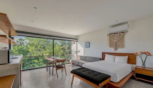 Relax in a serene bedroom with a large bed, a small table, and a sliding glass door leading to a balcony with a view of lush greenery.