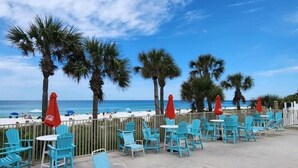 Une terrasse au bord de la plage, avec des chaises bleues et des parasols rouges, d’où l’on a une vue sur l’océan.